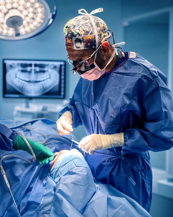 Sherif Kamel operating in theatre, gowned and gloved and wearing magnifying loupes, with a panoramic dental X-ray on the screen behind him.