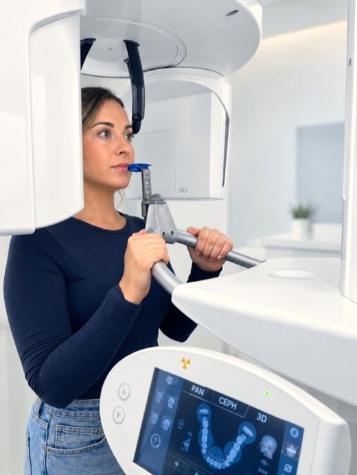 A patient positioned in a cone beam CT scanner, holding its handles, with the scanner's touchscreen in the foreground.
