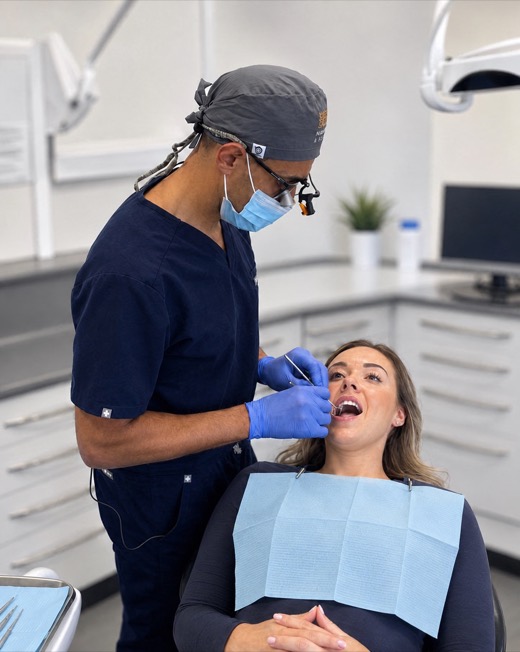 Sherif Kamel, wearing loupes and a surgical mask, examining a patient's teeth in a treatment room.